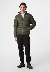 Vihreä toppatakki, jossa vetoketju edessä ja pystykaulus, koristeltu tikatulla kuviolla. Mustat housut ja urheilukengät täydentävät asun.