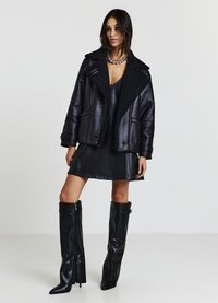 Giacca di pelle nera con colletto in shearling, abbinata a un vestito di pelle nera attillato e stivali neri alti con zip.