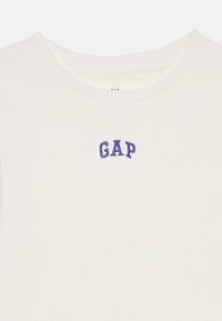 T-shirt à col rond blanc à côtes avec un petit logo "GAP" brodé en violet, centré sous le col.