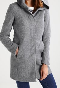 Een vrouw in een aansluitende grijze jas tot halverwege de bovenbenen met een verborgen sluiting aan de voorkant en haar handen in de zijzakken, gecombineerd met blauwe jeans.