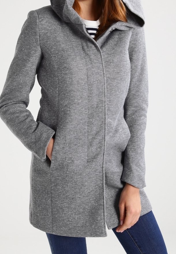 Een vrouw in een aansluitende grijze jas tot halverwege de bovenbenen met een verborgen sluiting aan de voorkant en haar handen in de zijzakken, gecombineerd met blauwe jeans.