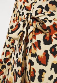 Tecido de estampa de leopardo em creme, laranja e preto. Inclui um detalhe de cinto amarrado com uma textura suave e caimento leve. Fecho detalhado com botão.