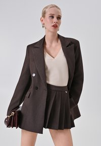 Giovane donna in blazer marrone e gonna plissettata, maglietta crema a scollo a V, che tiene una borsa marrone scuro, in piedi contro uno sfondo chiaro e neutro.
