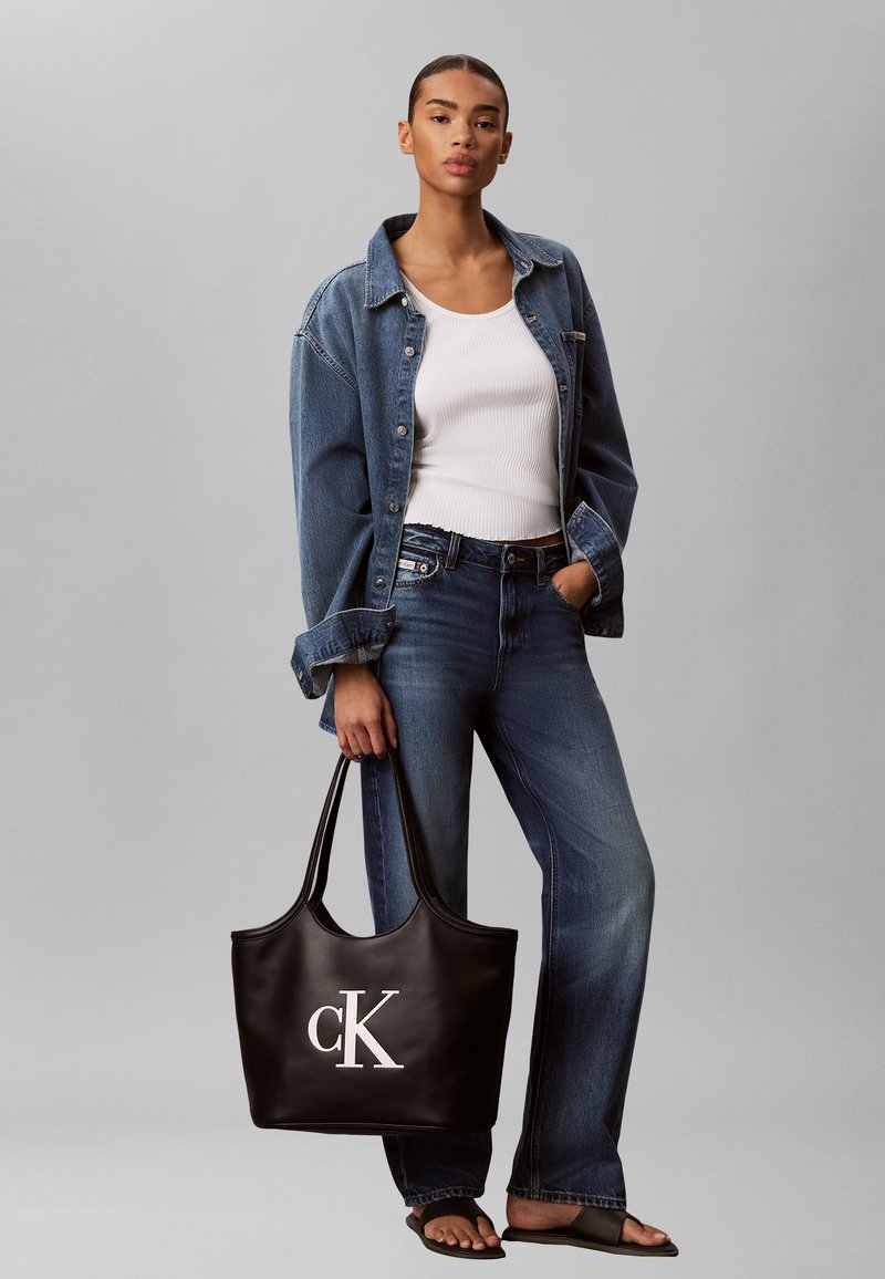 Femme portant une veste en jean et un jean avec un débardeur blanc, tenant un sac cabas noir Calvin Klein, debout devant un fond gris uni.