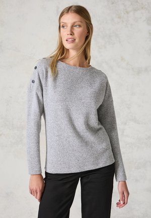 Pull en tricot gris avec un col bateau, présentant des accents de boutons sur l'épaule et des manches longues, associé à un pantalon noir.