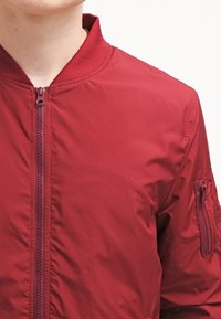 Burgunderfarbene Bomberjacke mit einem Reißverschluss vorne, geripptem Kragen und einer Reißverschlusstasche am Ärmel, mit einer glatten, glänzenden Textur.