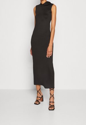 Femme portant une robe midi noire sans manches avec une capuche et des sandales noires à talons hauts à brides, debout sur un fond blanc.