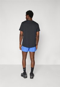 Schwarz T-Shirt mit kurzen Ärmeln kombiniert mit blauen Shorts, die einen lässigen Sitz zeigen. Das Modell trägt schwarze Socken und graue Sportschuhe.