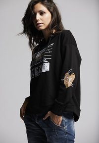 Black hoodie featuring a graphic design, long sleeves, and a side heart accent in beige. Text and patterns in white, relaxed fit.