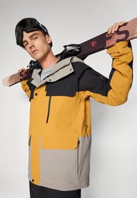 Lyžiarska bunda vo žltej, sivej a čiernej farbe s kapucňou, veľkými prednými vreckami a zipsami. Snowboard a okuliare sú držané cez rameno.