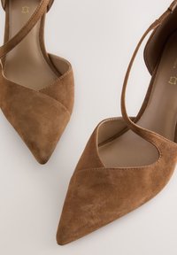 Rudi zomšiniai aukštakulniai batai su smailia nosimi, turintys kampuotą išpjovų dizainą ir susikertančias čiurnos dirželius. Lygi vidinė pamušalas.