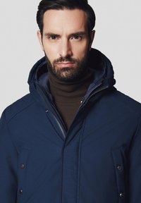 Granatowa kurtka z zapięciem na zamek i guzik, z kapturem i bocznymi kieszeniami, noszona na brązowym swetrze z golfem. Gładka tekstura.