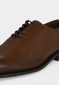 Καφέ δερμάτινο οξφορντ παπούτσι με κλειστό κορδόνι, ραμμένες λεπτομέρειες, στρογγυλεμένο μύτη και μαύρη σόλα, απεικονίζεται από μία γωνία μπροστινής πλευράς.