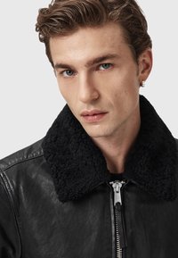 Sort læderbomberjakke med sort shearling-krave, sølv lynlåslukning og en blød tekstur. Fremstillet mod en ensfarvet baggrund.