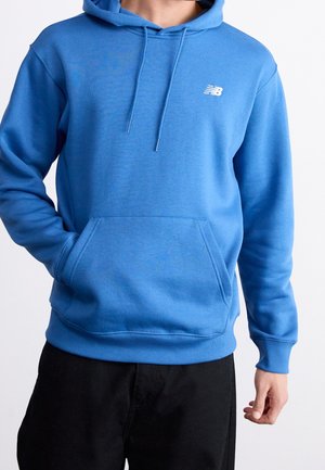 Mężczyzna w niebieskiej bluzie New Balance z kieszenią z przodu i czarnych spodniach, stojący na białym tle.