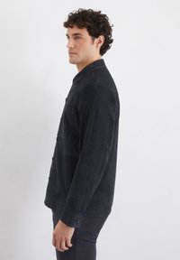 Schwarze Wildlederjacke mit Knopfleiste, spitzem Kragen, einer Brusttasche und langen Ärmeln. Glatte Textur mit figurbetonter Passform.