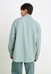 Man facing away wearing a loose, vertically striped green and white shirt with green cargo pants against a plain white background.