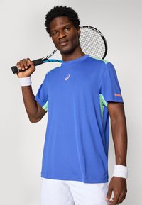 Young man wearing blue and mint sports shirt, white shorts, and wristbands holding a black and blue tennis racket over his shoulder.