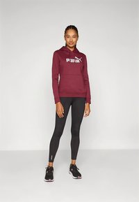 Camisola com capuz vinho com logótipo branco da Puma, bolso frontal; combinada com leggings pretas e ténis pretos com detalhes em rosa. Acabamento de tecido liso.