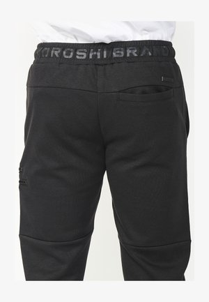 Pantalones de chándal negros hechos de un tejido suave con una cinturilla acanalada, que cuentan con un logotipo sutil, un bolsillo lateral y detalles de costura estructurada.