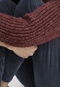 Personne portant un pull en maille bordeaux et un pantalon en velours côtelé bleu marine, assise avec les bras entourant ses genoux pliés.