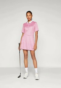 Φόρεμα γκολφ σε ανοιχτό ροζ χρώμα με λευκές ρίγες, κοντά μανίκια και γιακά. Διαθέτει εφαρμοστό μέση και φαρδύ φούστα; σε συνδυασμό με λευκά αθλητικά παπούτσια.