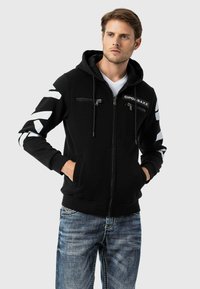 Schwarze Zip-Hoodie mit Kängurutaschen, weißen Chevron-Mustern an den Ärmeln und vorderen Reißverschlusstaschen; aus einem weichen, texturierten Stoff gefertigt.