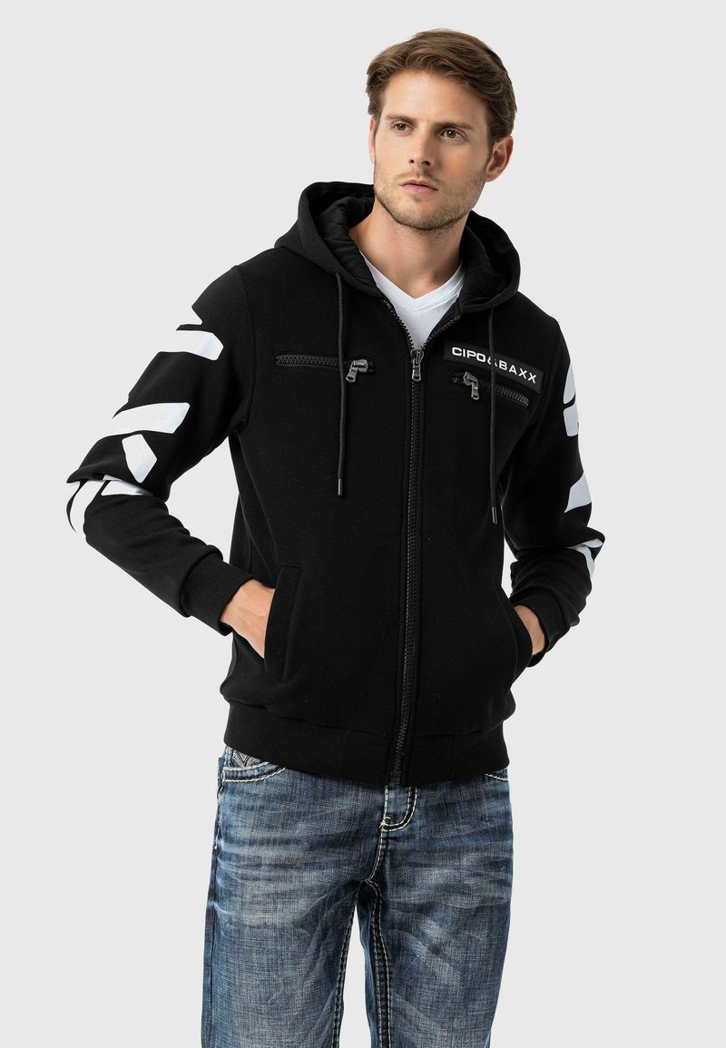 Schwarze Zip-Hoodie mit Kängurutaschen, weißen Chevron-Mustern an den Ärmeln und vorderen Reißverschlusstaschen; aus einem weichen, texturierten Stoff gefertigt.