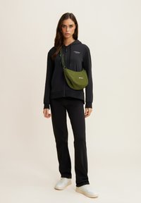 Sudadera negra con cremallera y logo, combinada con pantalones negros y zapatillas blancas. Detalle adicional: bolsa cruzada verde con correa ajustable.