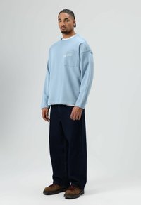 Lichtblauwe sweater met ronde hals en een zak aan de voorkant, met witte geborduurde tekst, gecombineerd met donkere wijde jeans en bruine wandelschoenen.