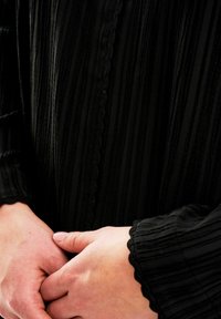 Schwarz strukturiertes Stoffkleidungsstück mit vertikalen Falten, gewellten Kanten und langen Ärmeln, die Hände am Taillenbereich gefaltet, minimale sichtbare Details.