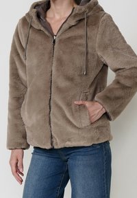 Personne portant un sweat à capuche zippé duveteux taupe avec cordons, main dans la poche, associé à un jean bleu sur un fond uni.