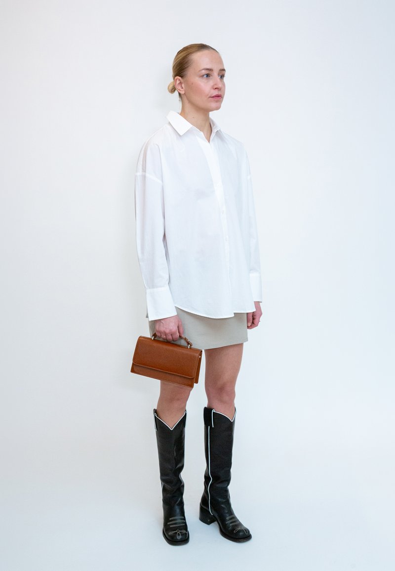 Femme debout sur fond blanc, portant une chemise blanche, une jupe beige, des bottes noires montant jusqu'aux genoux, et tenant un petit sac à main marron.