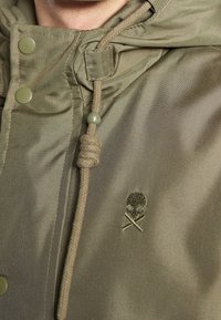 Olivengrøn jakke med tekstureret stof, der har en snoretrækshætte, metalsnaps og et lille broderet skelet og korslagte knogler logo.