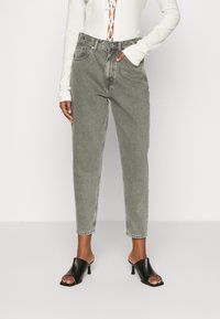 Modelo vistiendo jeans de talle alto grises y sandalias de tacón abiertas negras, de pie contra un fondo blanco liso.