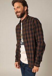 Bruine en zwarte geblokte overhemd, lange mouwen, knoopsluiting aan de voorkant, enkele borstzak, katoen stof, gedragen over een lichte graphic T-shirt.