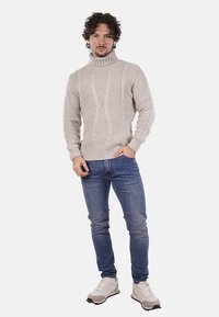 Maglione beige a maglia con collo alto, caratterizzato da un motivo a treccia. Abbinato a jeans blu e sneaker chiare.