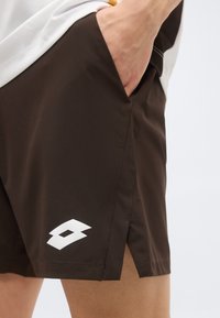Κοντινό πλάνο ενός ατόμου που φορά σκούρα καφέ αθλητικά σορτς με λευκό λογότυπο, με το χέρι τοποθετημένο άνετα στην πλαϊνή τσέπη.