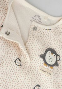 Bawełniany strój niemowlęcy z miękkiej beżowej tkaniny, białymi kropkami i haftowanymi detalami w postaci pingwina. Posiada zapięcia na napy oraz metkę "little ones".