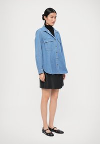 Hellblaues, oversized Denim-Hemd mit zwei Brusttaschen, braunen Knöpfen, kombiniert mit einem schwarzen Lederröck und schwarzen Mary Jane Schuhen.