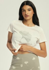 White cotton t-shirt featuring a detailed ginkgo leaf print, styled with a rounded neckline. Paired with olive green pants with matching leaf designs.