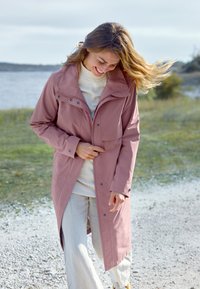 Ljusrosa vattentät jacka med hög krage, tryckknappar och en framficka, ihop med ljusa byxor, i en utomhusmiljö.