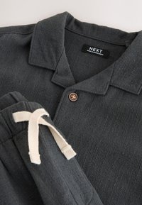 Camisa texturizada gris oscuro con cuello y botón, combinada con pantalones a juego con cordones blancos.