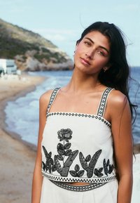 Top court blanc avec broderie florale noire, décolleté carré et bretelles à motif. Porté dans un cadre de plage avec des vagues en arrière-plan.