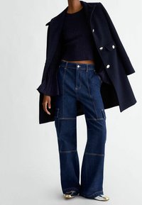 Mannequin portant un pull côtelé bleu foncé, un manteau assorti posé sur les épaules, un pantalon en denim patchwork foncé et des chaussures plates à bout métallique.