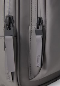 Mochila gris con acabado mate, tela impermeable, cremalleras dobles con tiradores negros y detalles de costura discretos.