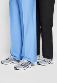 Två personer står sida vid sida i grå och vita sneakers, den ena i ljusblå vida byxor och den andra i svarta smala byxor.