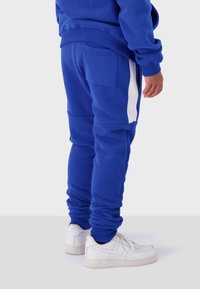 Blue fleece joggers with side white stripes, ribbed cuffs, and a back pocket, paired with white sneakers. Soft texture and fitted design.