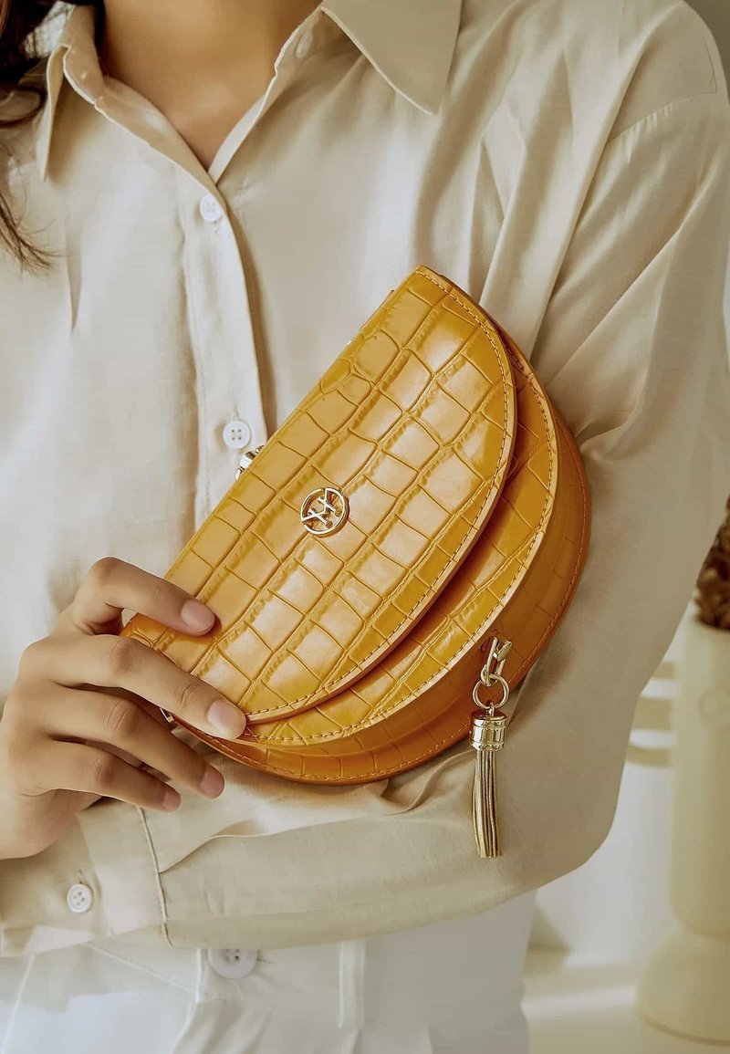 Personne en chemise beige clair tenant un petit sac à main moutarde texturé avec une fermeture dorée et un détail pompon.