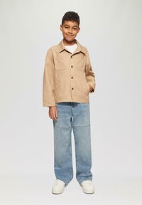 Beżowa bawełniana kurtka z dwoma przednimi kieszeniami, zapinana na guziki, z białymi guzikami, w zestawieniu z jasnoniebieskimi, oversize'owymi dżinsami i białymi sneakersami.